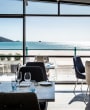 Dining room with upholstered chairs and wooden tables in the foreground, and a floor-to-ceiling panoramic window looking out to a sandy beach and blue sea