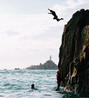 A man leaping into the water from the the top of larger rock with Corbiere Lighthouse in background.