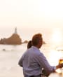 A couple sit looking out to La Corbiere Lighthouse at sunset having a drink at Corbiere Phare