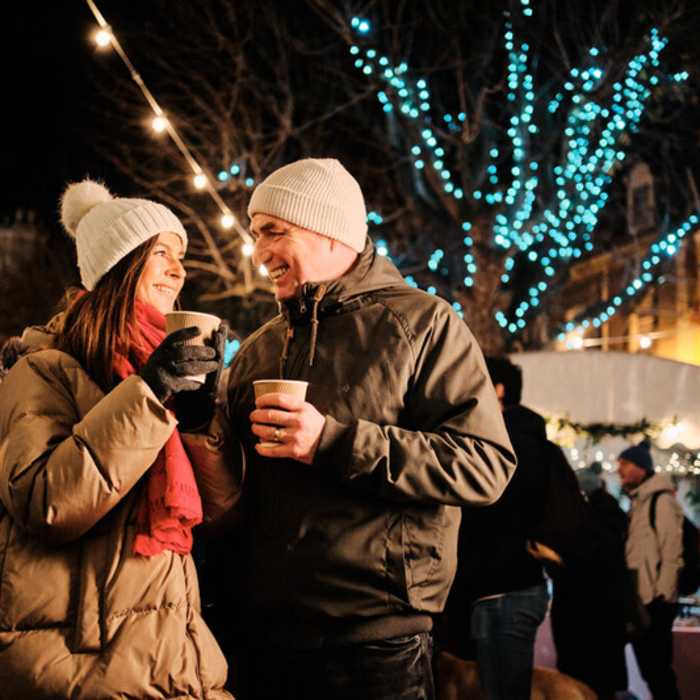 Couple smiling each other. They are wearing warm, wooly, coats are are surrounded by people and bright Christmas lights.