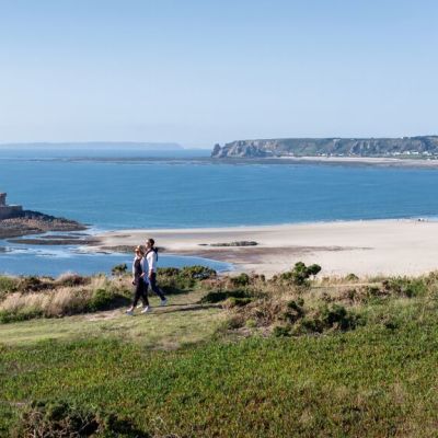 Couple walking on a grassy headland overlooking a sandy beach and blue seas